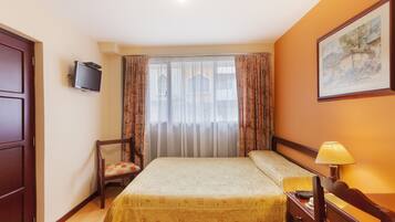A hotel room with a bed, a chair, a desk with a lamp, a television, and a painting on the wall.
