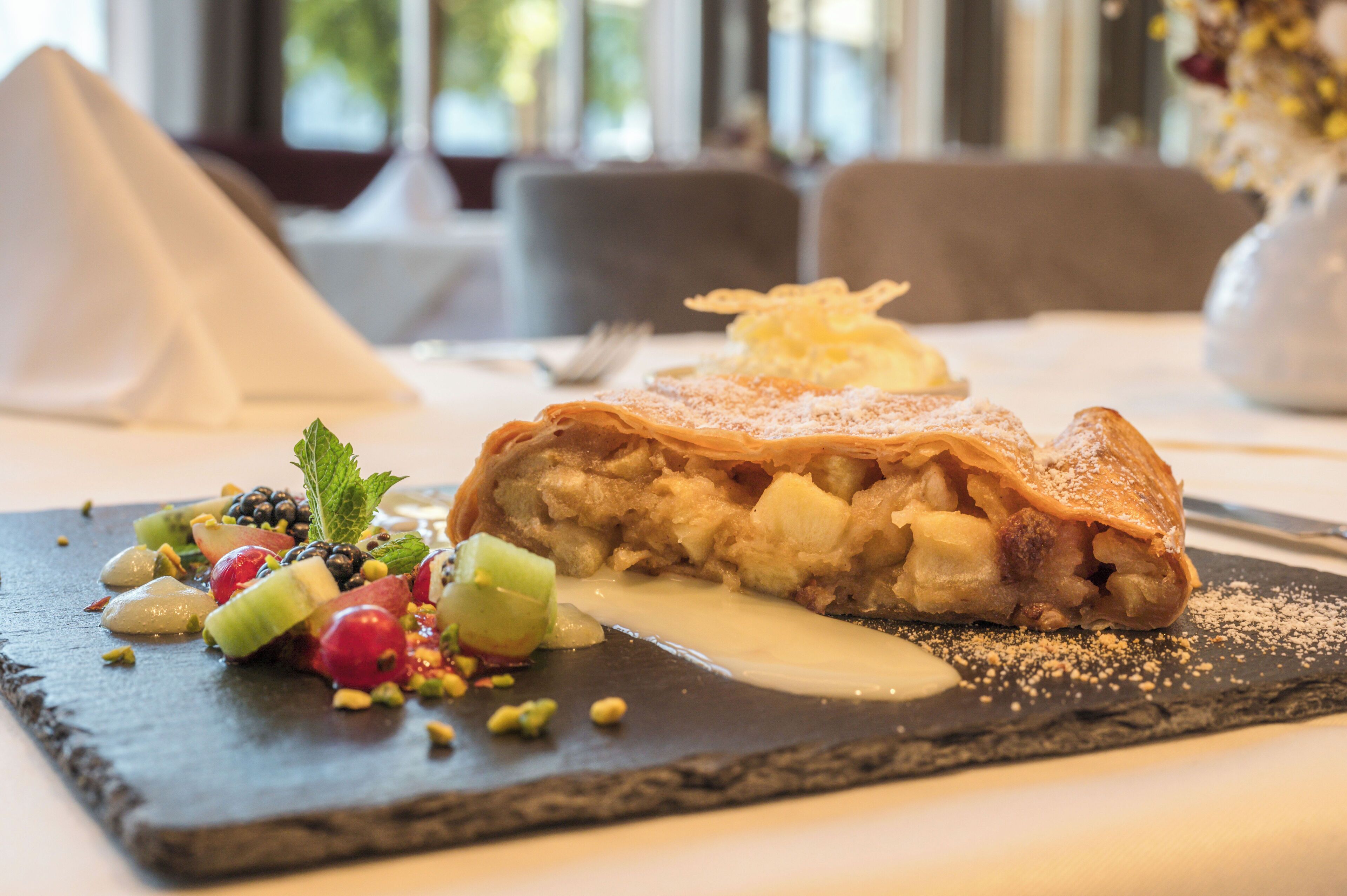 Un postre sobre un plato de pizarra con frutas y nueces, un objeto en forma de pirámide y un jarrón con flores al fondo.