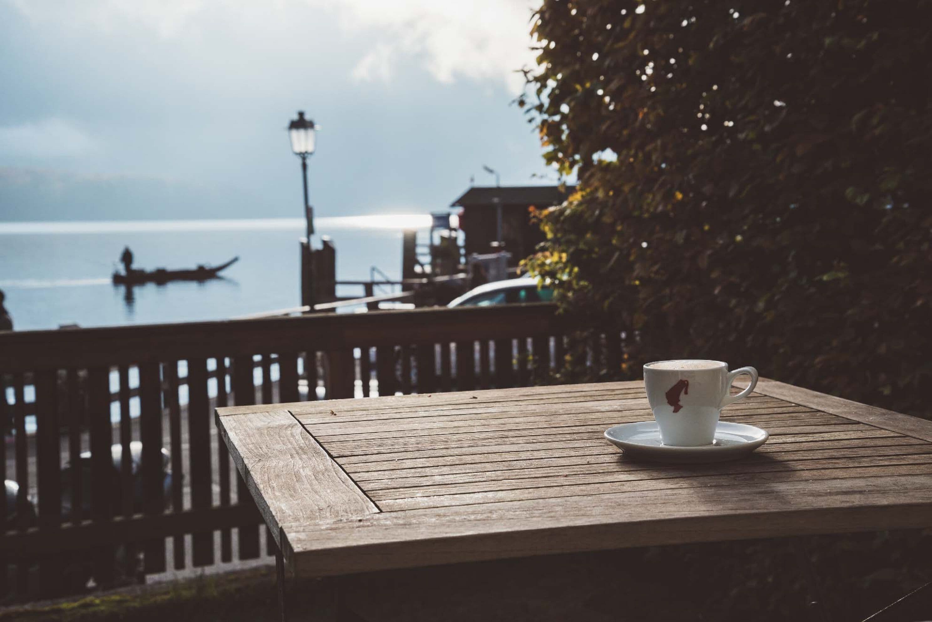 Una mesa de madera con una taza y platillo blancos, con vista a un cuerpo de agua con un bote y una costa lejana.