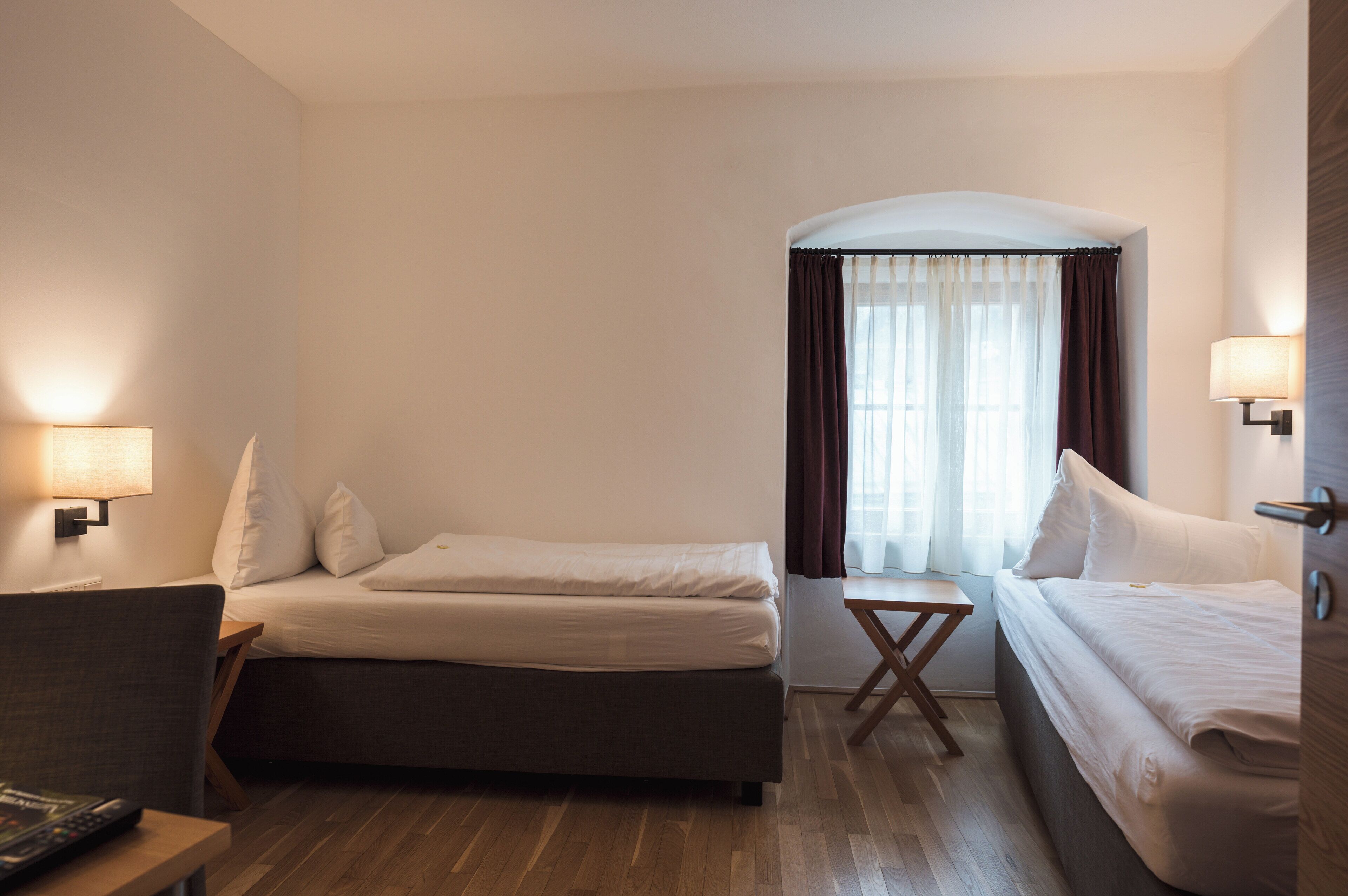 A hotel room with two beds, a small table, and a chair. There is a window with curtains and a lamp on the wall.
