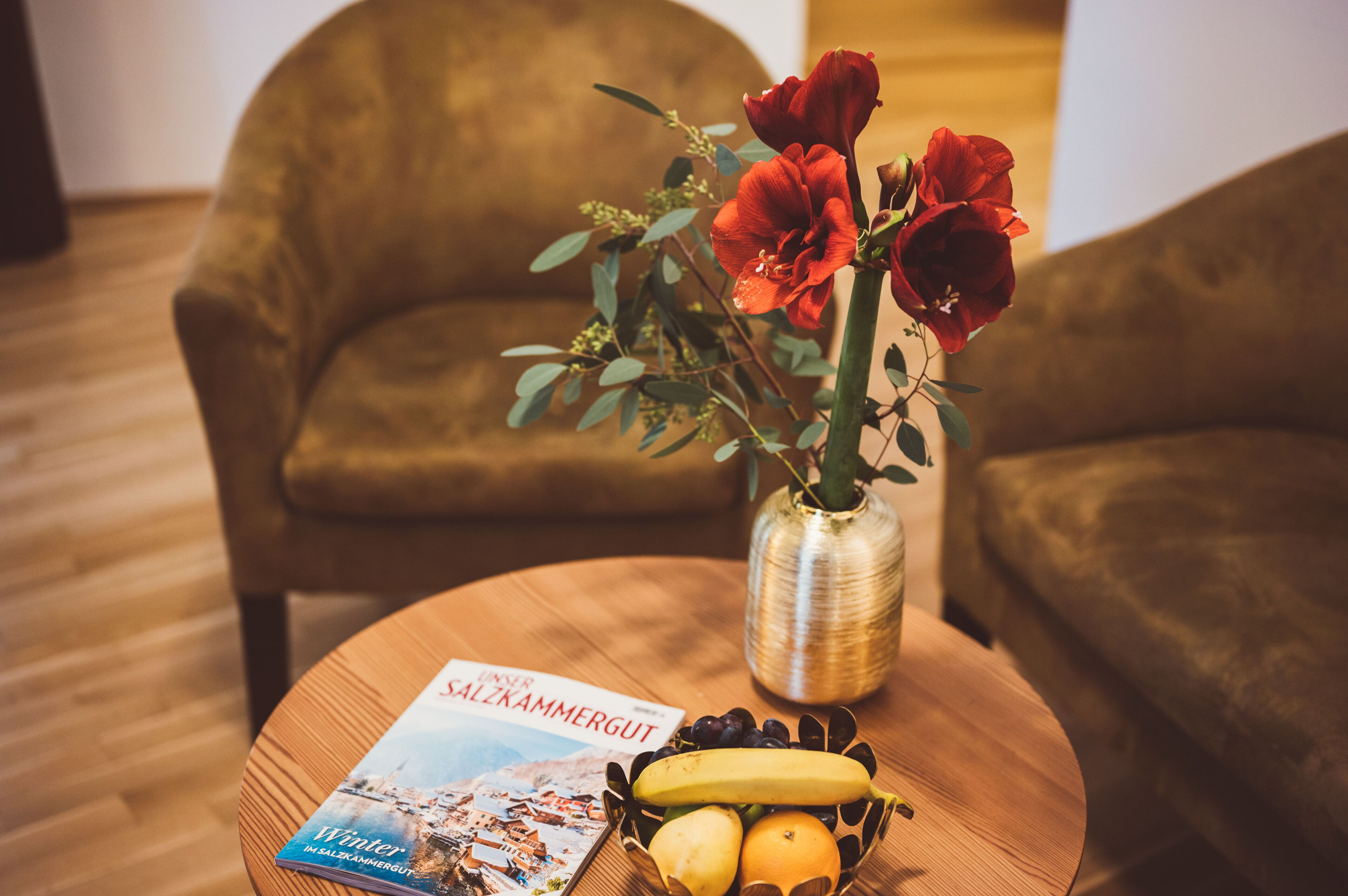 A round wooden table with a magazine, a vase with red flowers, and a bowl of fruit.