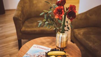 A round wooden table with a magazine, a vase with red flowers, and a bowl of fruit.