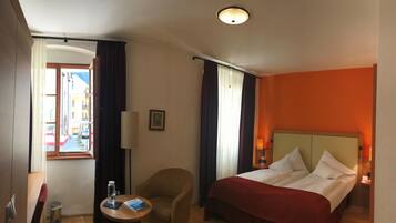 A hotel room with a bed, a chair, a small table with a book and a bottle, and a window with curtains.