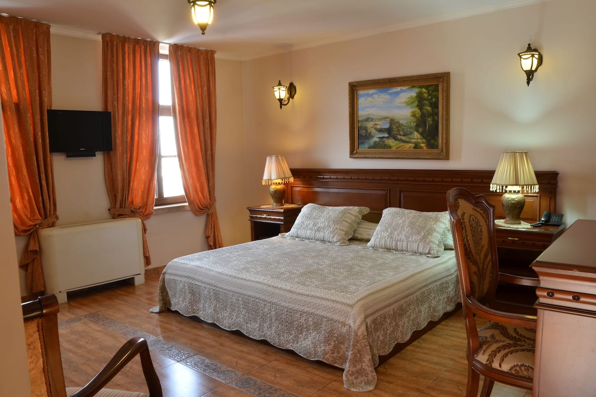 A neatly arranged bedroom with a large bed, two bedside tables, a dresser, a painting on the wall, and a radiator.