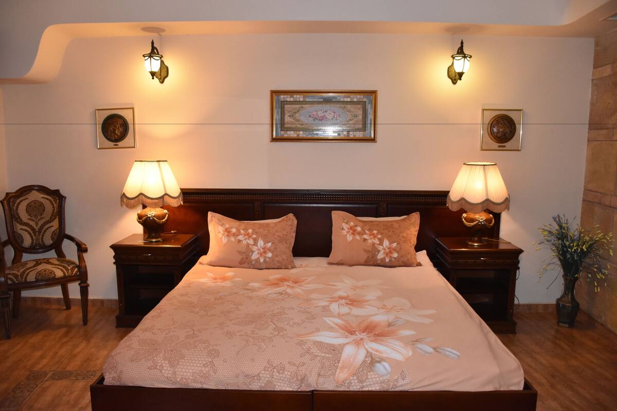 A neatly arranged bedroom with a large bed, two bedside tables with lamps, a chair, and framed pictures on the wall.