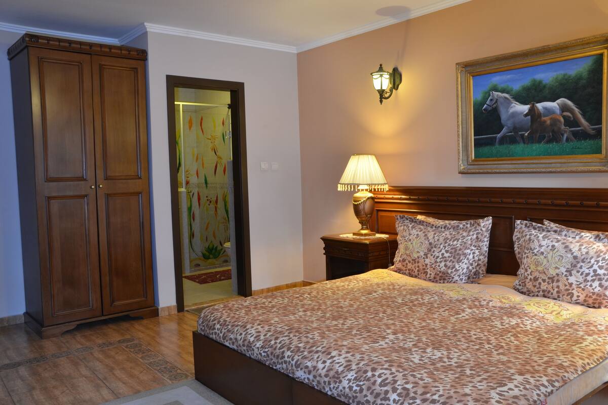 A bedroom with a large bed, a nightstand, a lamp, a wardrobe, and a painting on the wall.