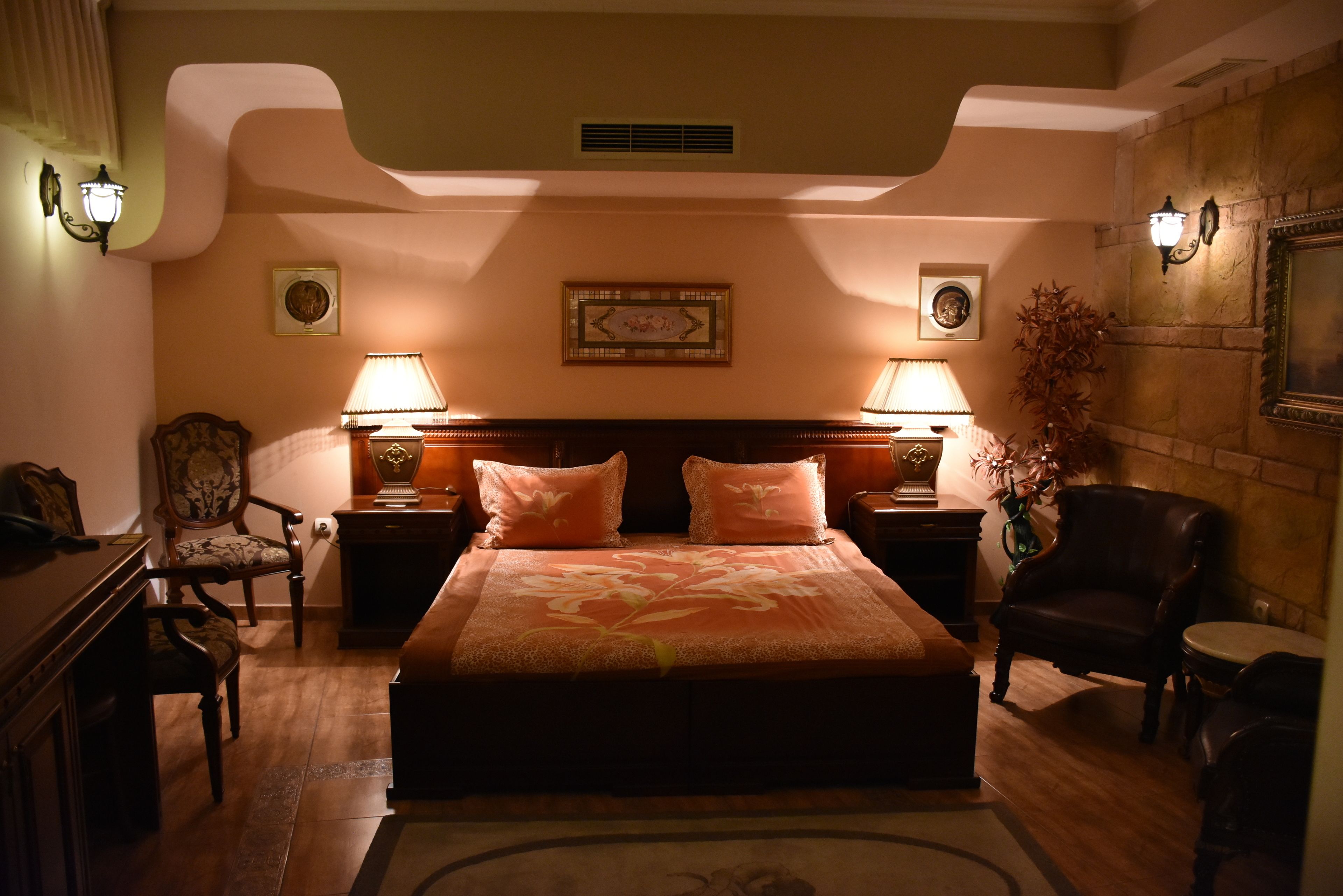 A hotel room with a large bed, two nightstands with lamps, a wooden dresser, a chair, and a potted plant.
