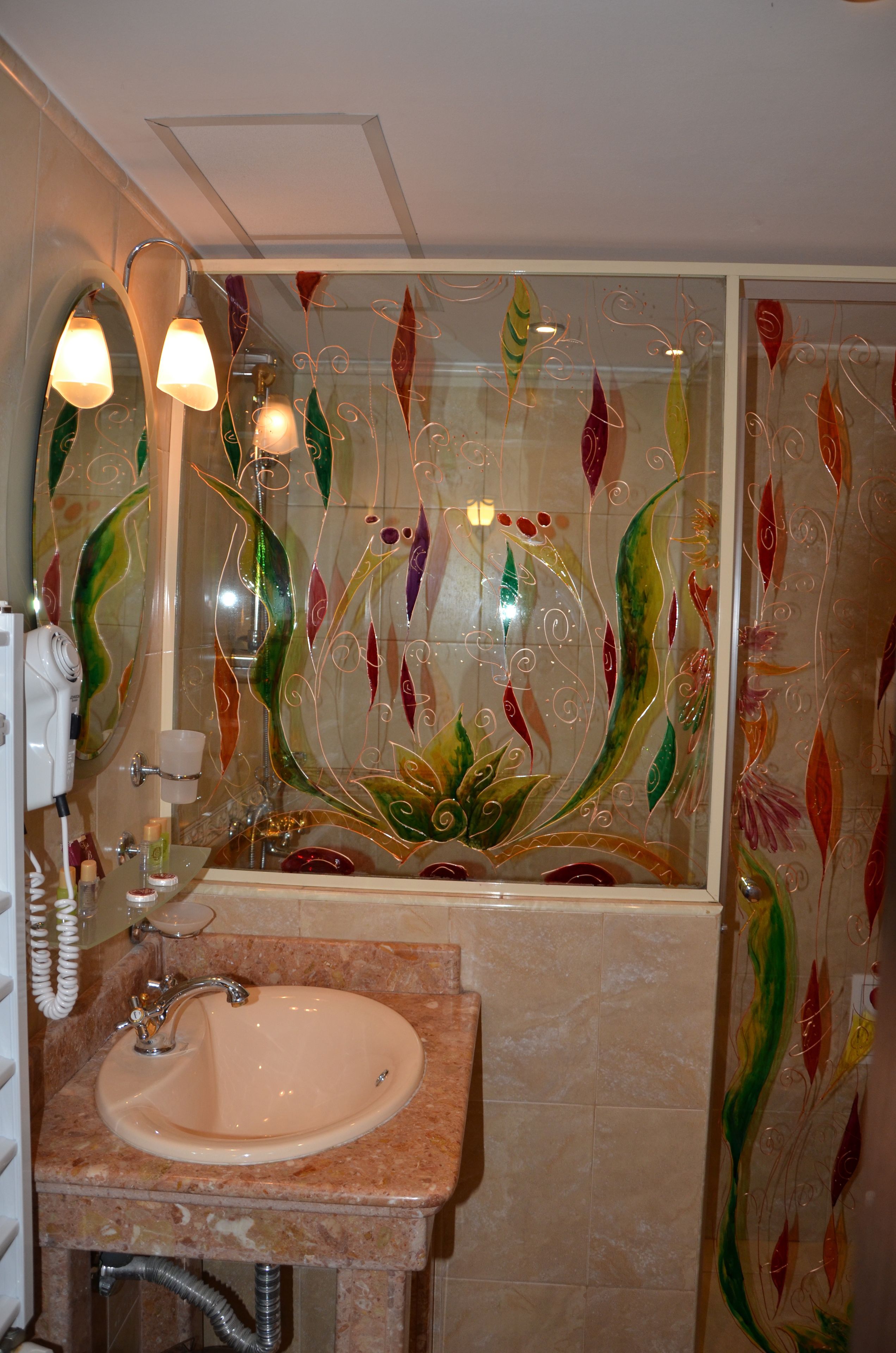 Une salle de bain dotée d’un lavabo en marbre, d’une cabine de douche en verre et d’un miroir orné d’un motif décoratif en forme de feuilles.