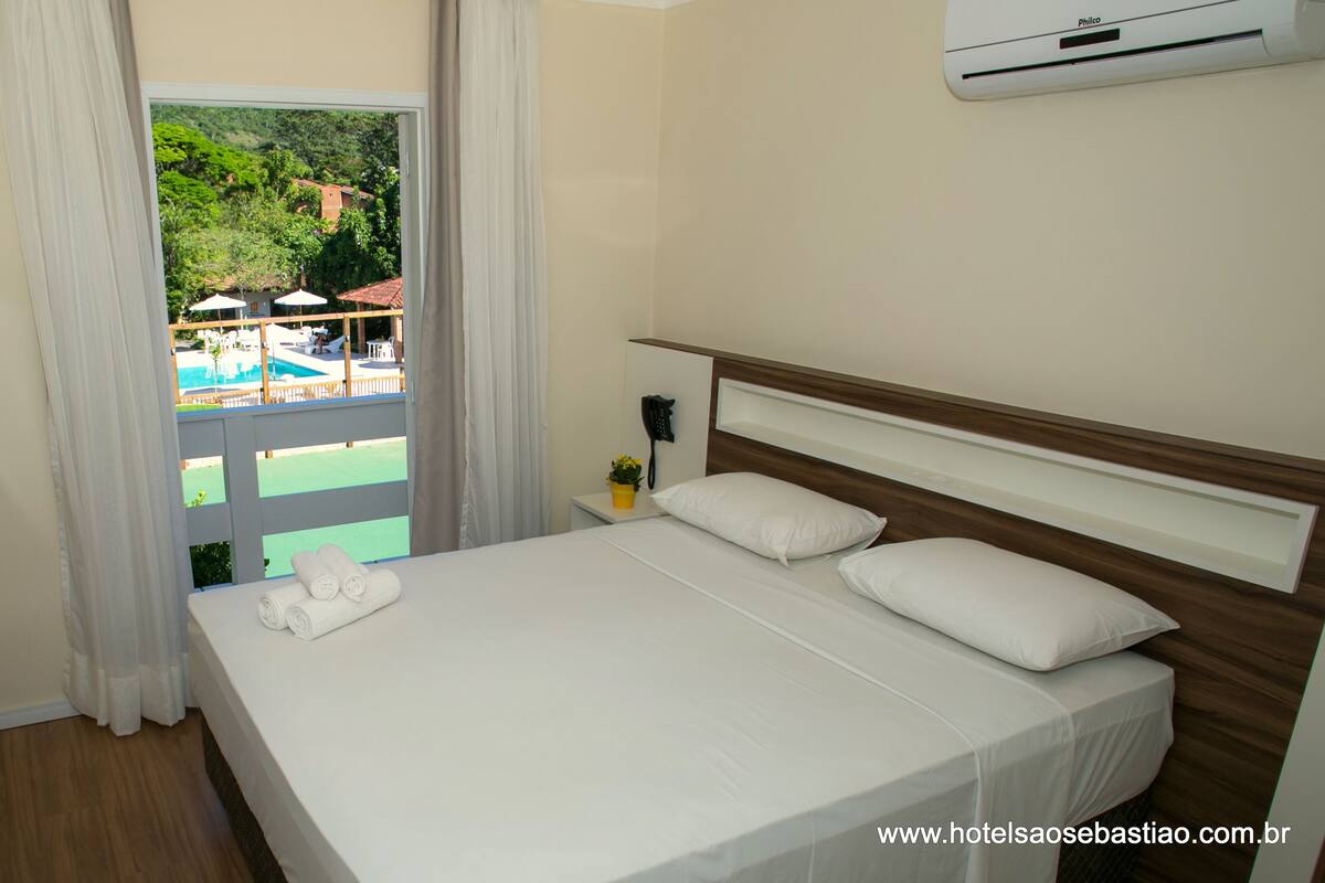 A hotel room with a bed, a TV, a potted plant, and a view of a pool and greenery outside.