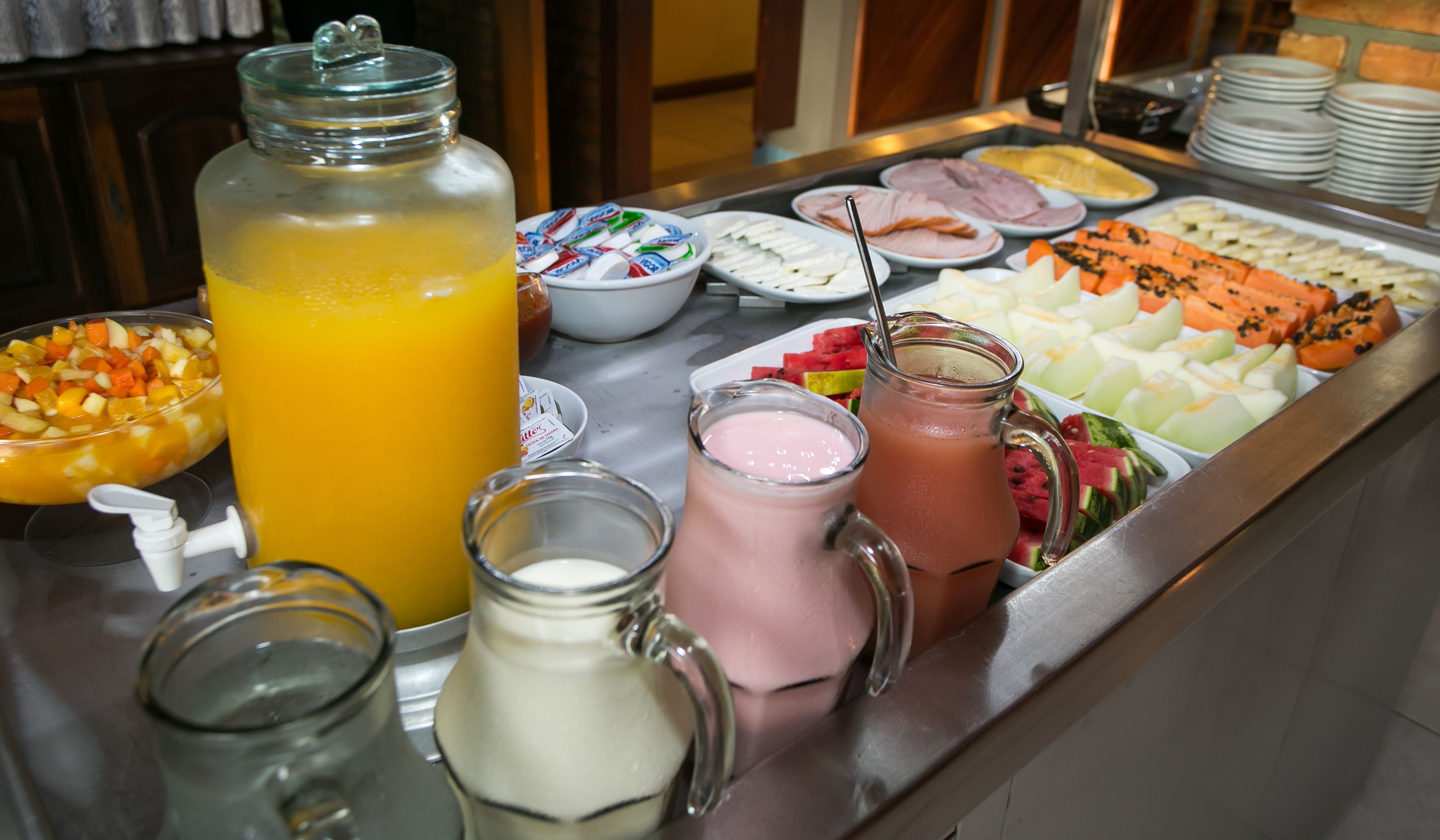 A buffet with various food items and beverages, including a large glass jar of orange juice, bowls of fruit, and a pitcher of pink smoothie.