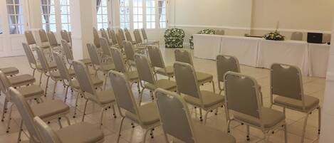 A conference room with rows of beige chairs, a white table with a laptop, and large windows with curtains.