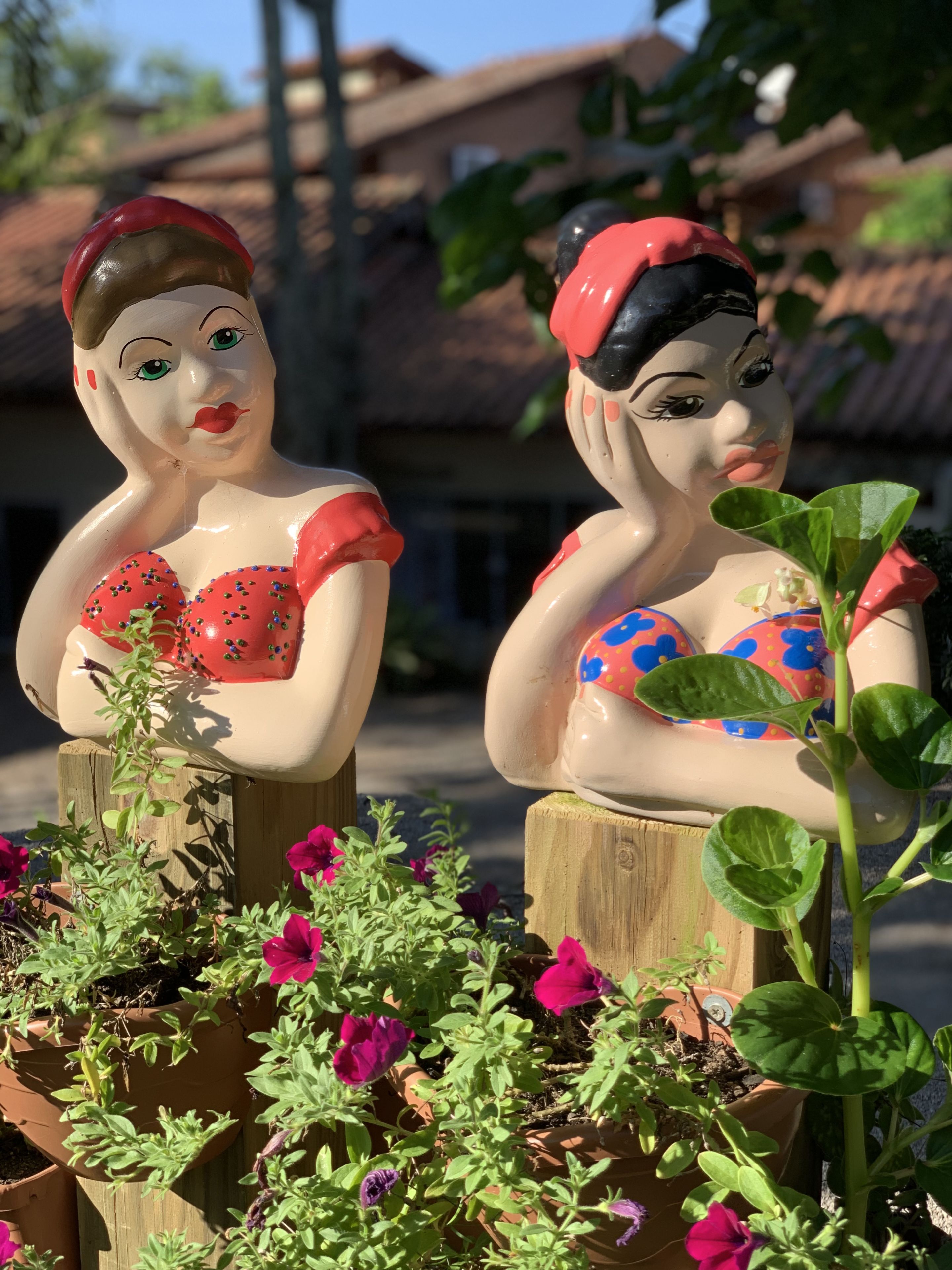 Two ceramic figurines of women in colorful attire, placed on wooden posts amidst potted plants with flowers.