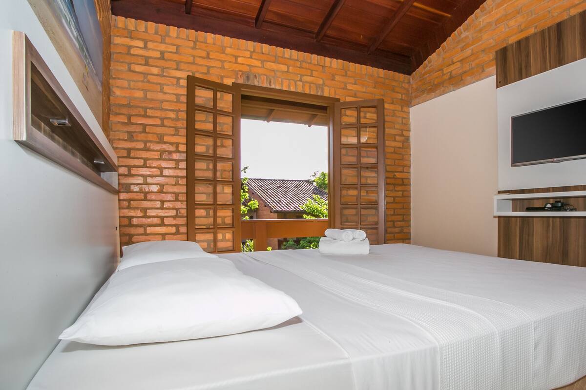 A hotel room with a brick wall, a wooden ceiling, a bed with white bedding, a TV mounted on the wall, and a window with a view of a building outside.