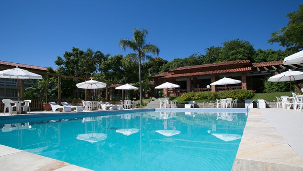A swimming pool with lounge chairs and umbrellas, surrounded by greenery and a building with a tiled roof. - Hotel Sao Sebastiao Da Praia (Florianópolis)