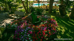 A garden with a fountain, colorful flowers, and a pool in the background. - Hotel Sao Sebastiao Da Praia (Florianópolis)