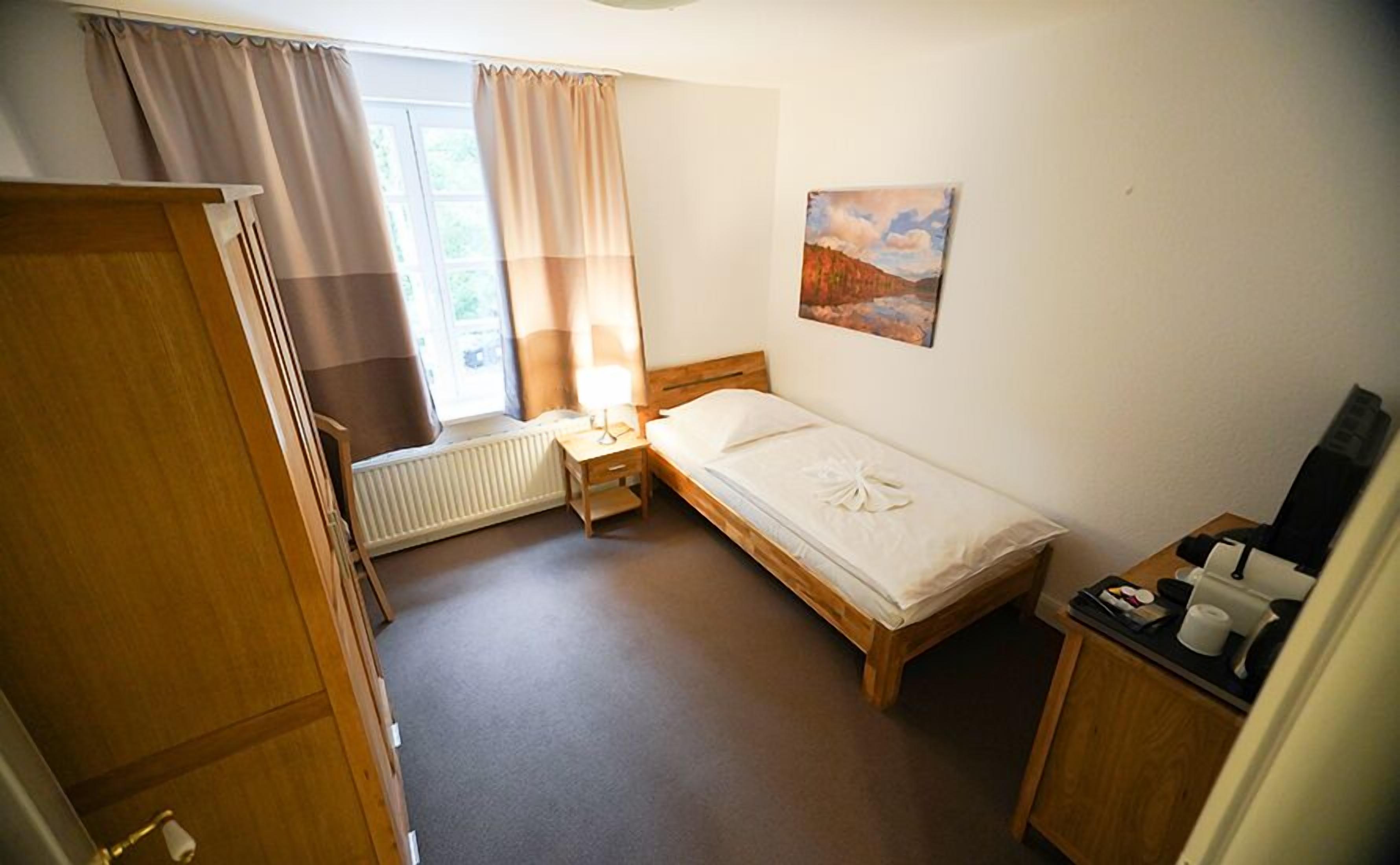 A small, neatly arranged bedroom with a single bed, a bedside table with a lamp, a radiator, a window with curtains, and a painting on the wall.