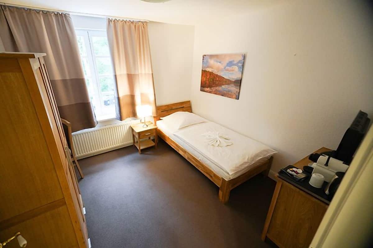 A small, neatly arranged bedroom with a single bed, a bedside table with a lamp, a radiator, a window with curtains, and a painting on the wall.