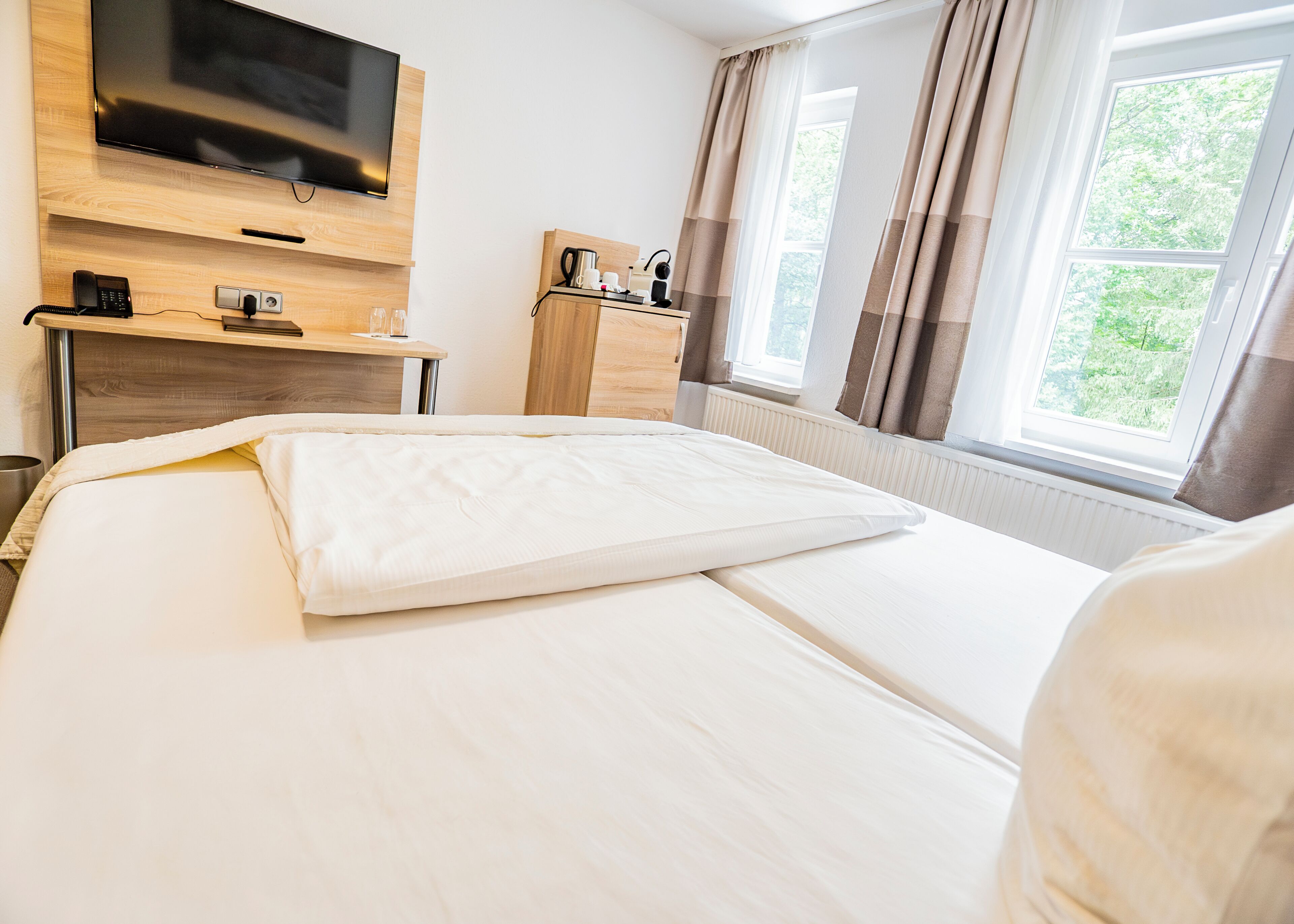 Ein Hotelzimmer mit Bett, Wandfernseher, Schreibtisch mit Kaffeemaschine und einem Vorhangfenster.