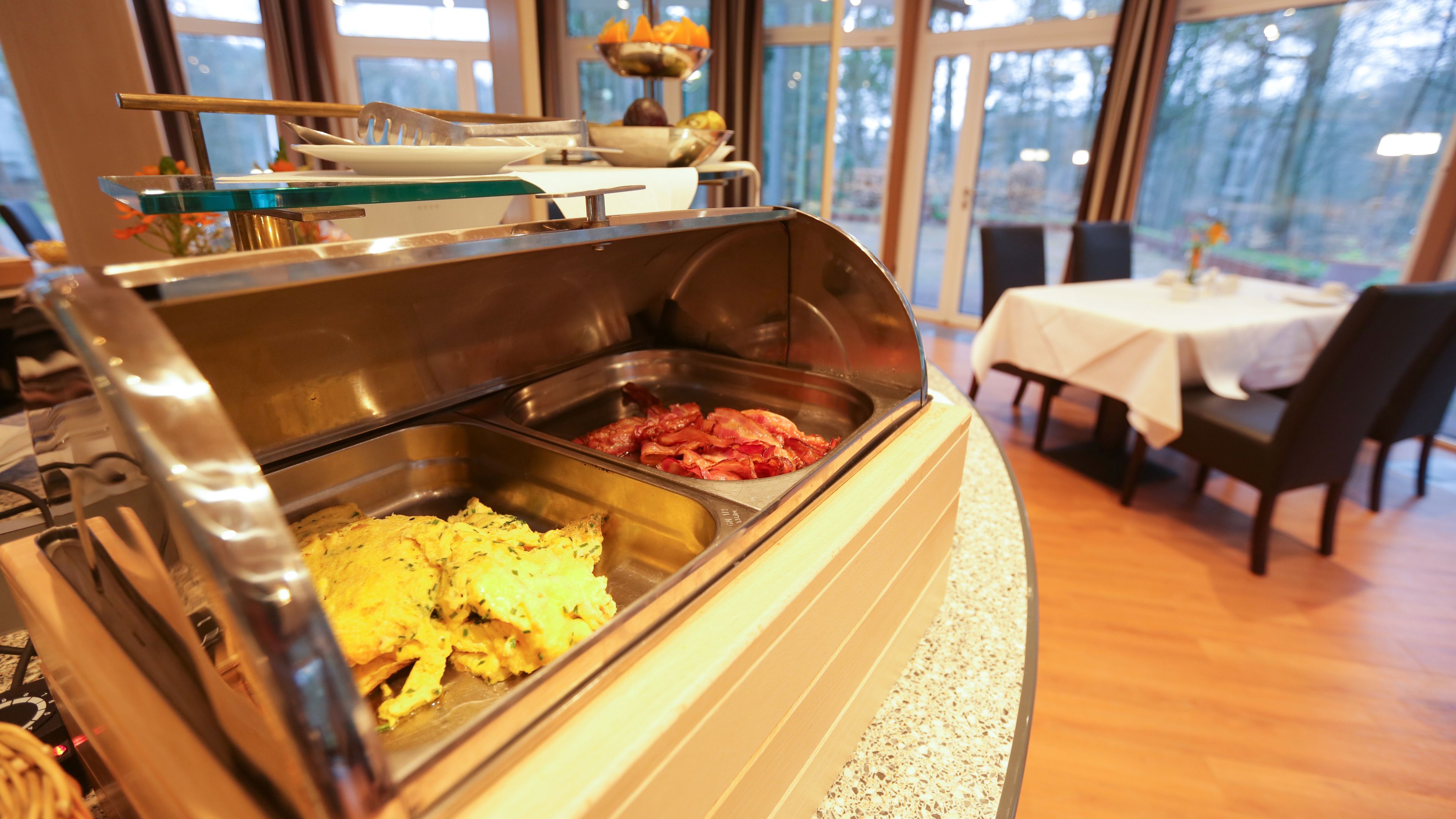 Zona de bufé con diversas opciones de comida, como huevos revueltos y tocino, en una habitación bien iluminada con vistas a árboles a través de amplios ventanales.