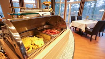 Zona de bufé con diversas opciones de comida, como huevos revueltos y tocino, en una habitación bien iluminada con vistas a árboles a través de amplios ventanales.