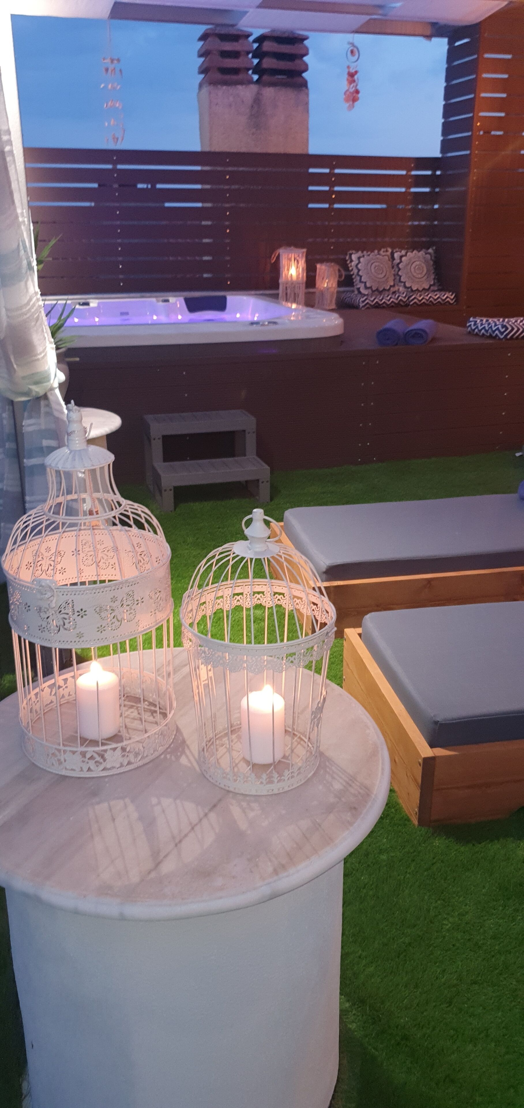 Un patio con dos faroles blancos estilo jaula para pájaros, una mesita y un banco de madera.