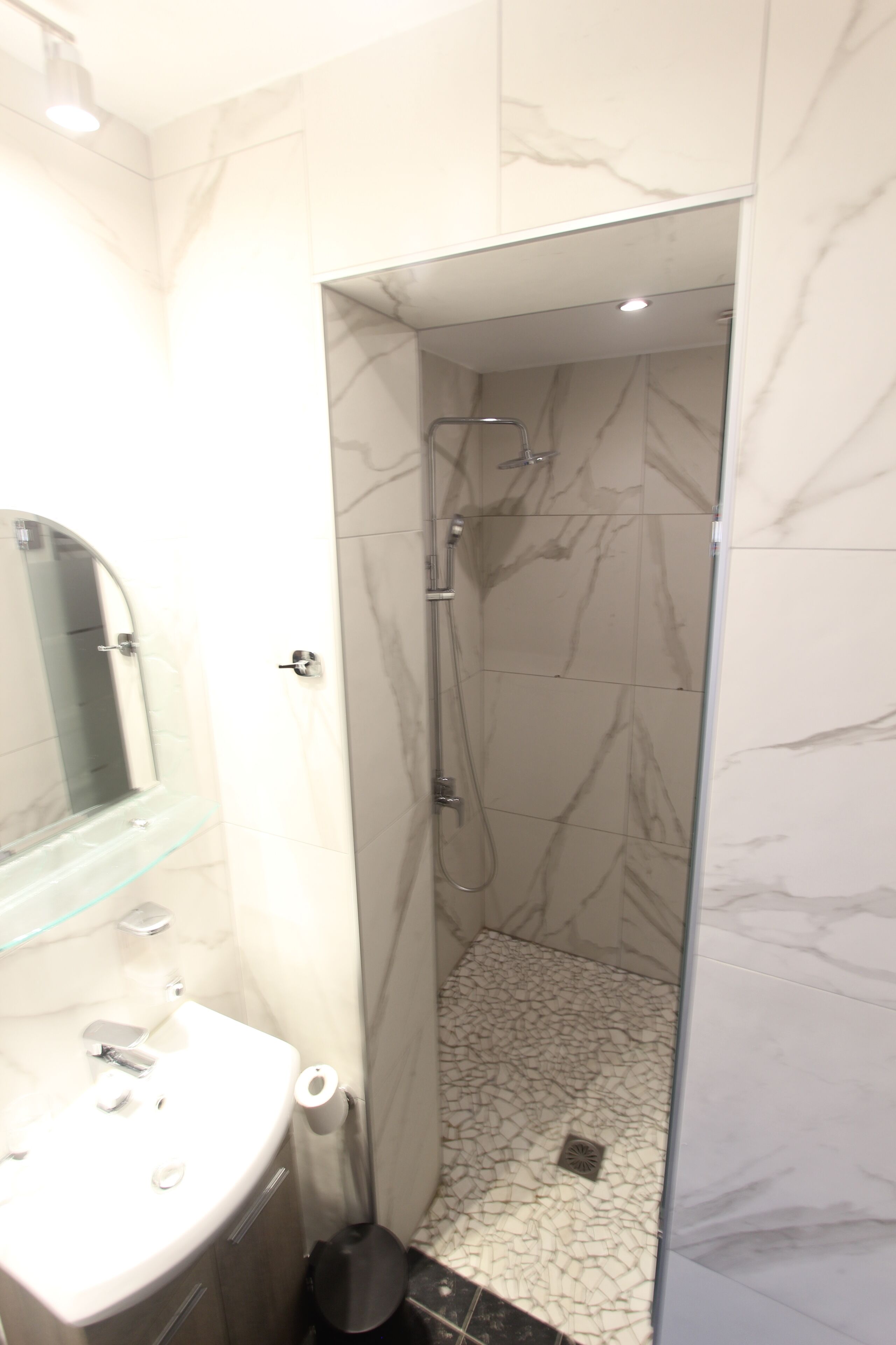 A bathroom with a glass shower enclosure, a sink with a glass shelf, and a marble-tiled floor.