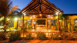 Una entrada de hotel con techo de paja, palmeras y fachada bien iluminada.