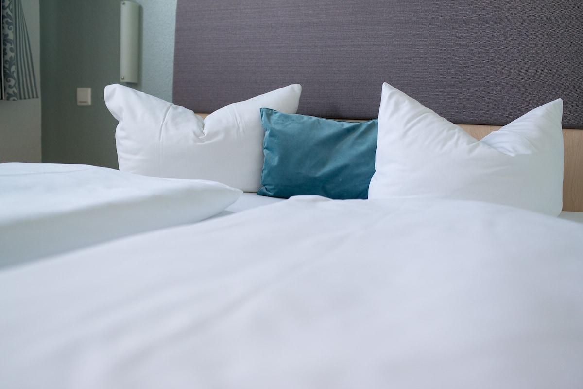 A neatly made bed with white and teal pillows, a wooden headboard, and a plain wall with a light switch.