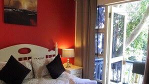 A bedroom with a white headboard, a bedside lamp, a red wall, and a window with a view of trees.
