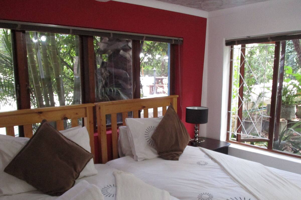 Un dormitorio con cabecera de madera, cama con ropa de cama blanca y almohadas marrones, mesita de noche con lámpara, y ventanas amplias con vistas a la vegetación.