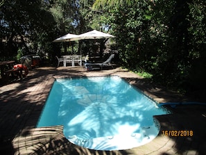 A rectangular swimming pool with a tiled edge, surrounded by a brick patio. There are lounge chairs and a table with an umbrella in the background. - Knysna Herons Guest House (Knysna)
