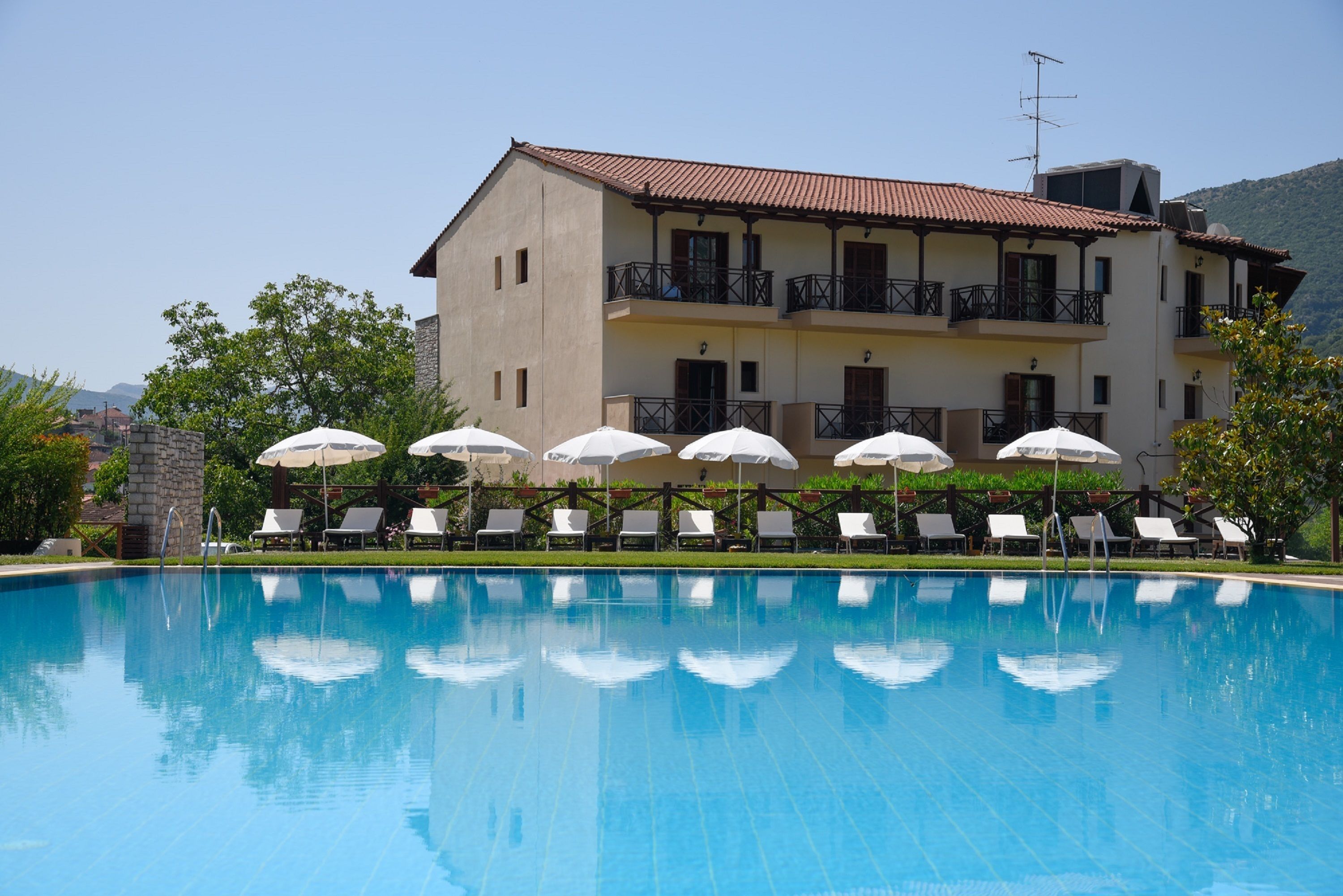Una piscina con sillones de descanso y sombrillas, un edificio con balcones y montañas al fondo.
