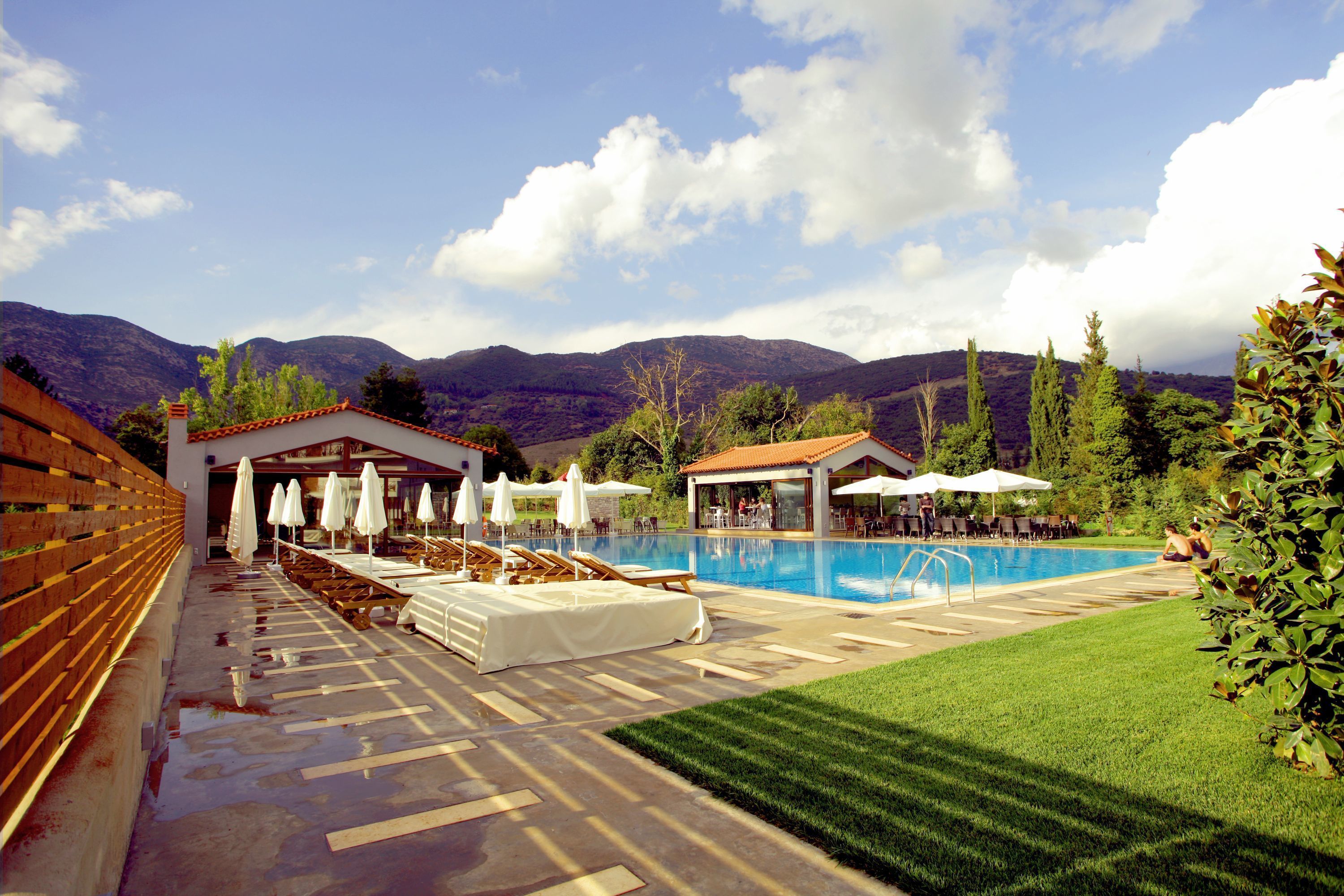 Un área de piscina con sillones de playa y sombrillas, una zona de estar cubierta y vistas a montañas al fondo.