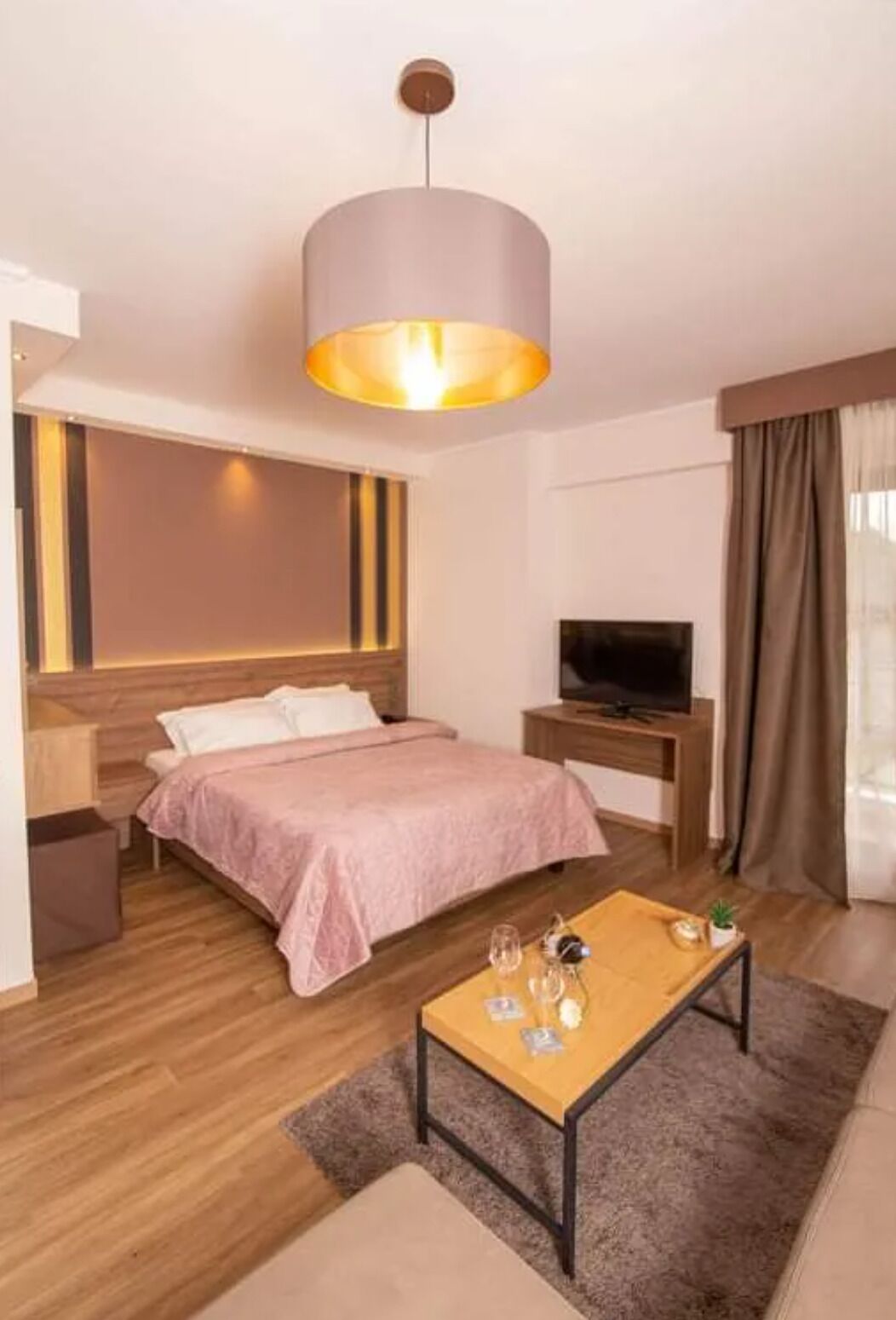 Habitación de hotel con cama, televisor, una mesita de madera y un amplio ventanal con cortinas.