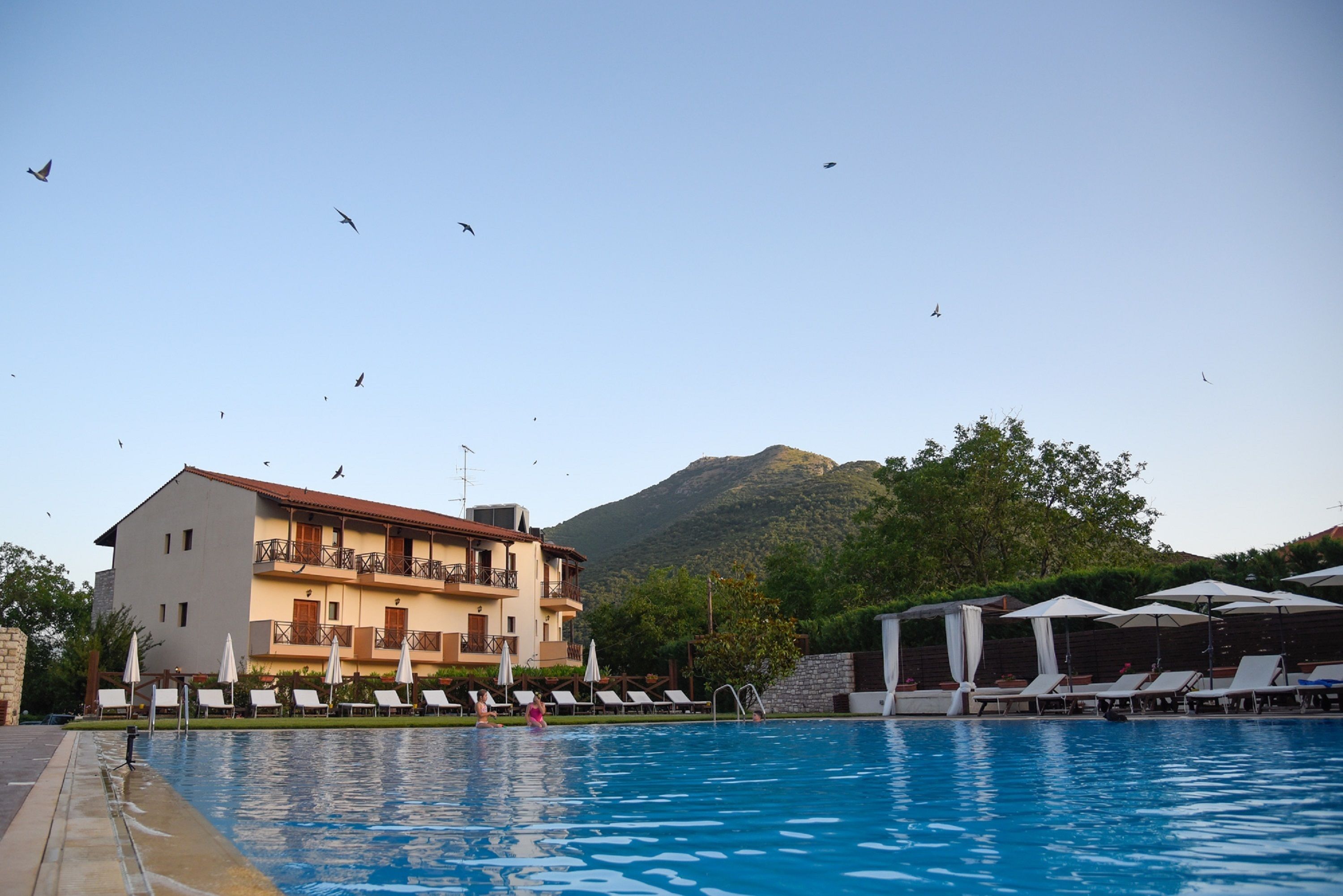 Una piscina con sillas de descanso y sombrillas, un edificio con balcones, y una montaña al fondo.