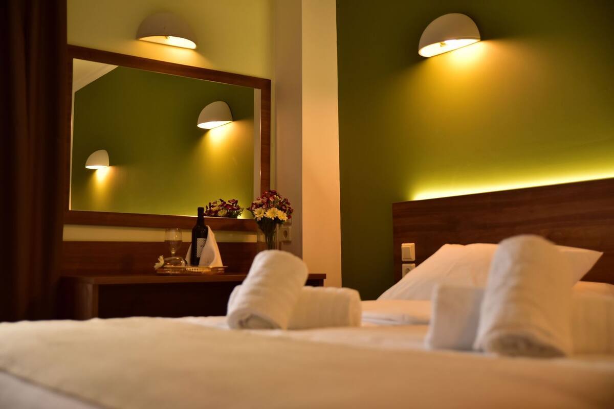 A hotel room with a bed, a nightstand with a bottle and glasses, a mirror, and a wall-mounted light fixture.