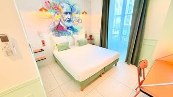 A hotel room with a bed, a bedside table, a chair, a window with curtains, and a wall with a colorful mural.