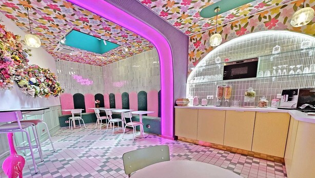 Een café met een geruite vloer, roze en witte stoelen, een roze ventilator en een plafond met bloemen.