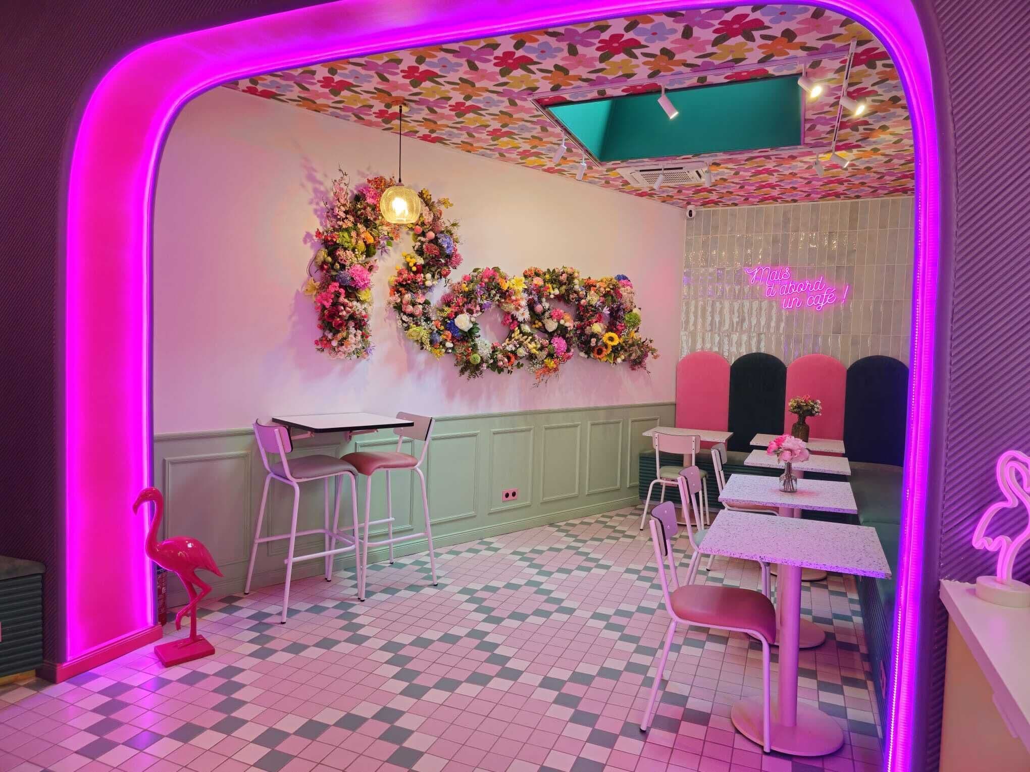 Ein Café-Interieur mit Schachbrettfußboden, rosa-weißen Stühlen, einem Neonlichtschild und floralen Wanddekorationen.