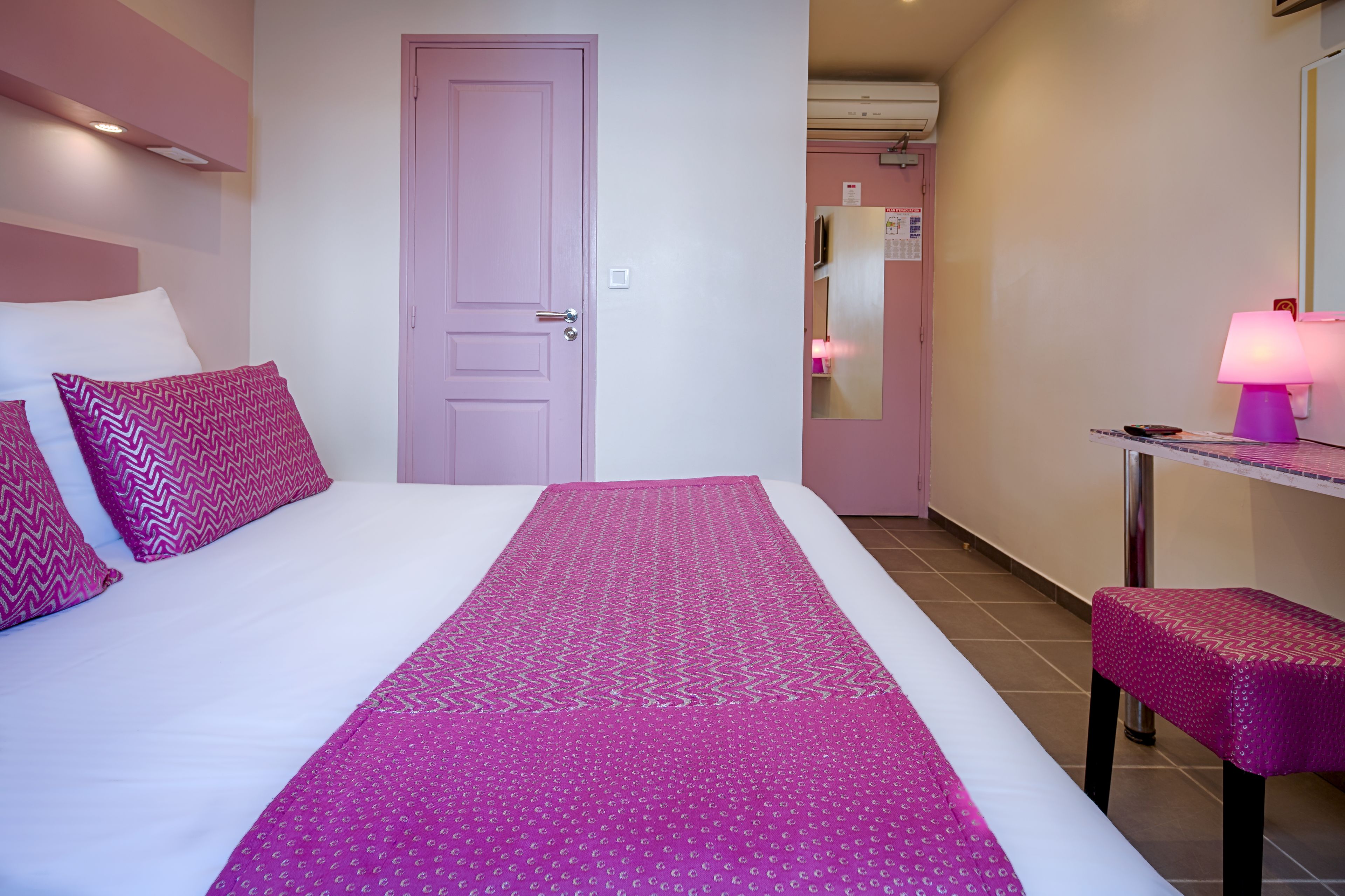 A hotel room with a bed, a pink door, a small table with a lamp, and a bench.