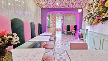 Interior de restaurante com chão quadriculado, assentos acolchoados em rosa e verde e decoração floral na parede.