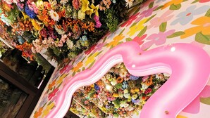 Een kleurrijke bloemenarrangement aan het plafond en een roze, golvende spiegel aan de muur.