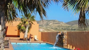 A swimming pool with a waterfall feature, surrounded by palm trees and a stone wall. - Hotel de Naturaleza Rodalquilar Spa Cabo de Gata (Nijar)