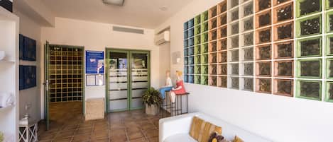 Un área de recepción con suelo de baldosas, un sofá blanco con cojín estampado, una pared con paneles de vidrio decorativos y una puerta verde que da a un área de spa.
