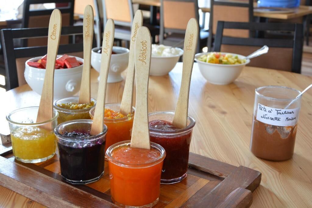 Una bandeja de madera con varios frascos de conservas y un frasco de salsa, cada uno con una cuchara de madera que lleva nombres escritos.