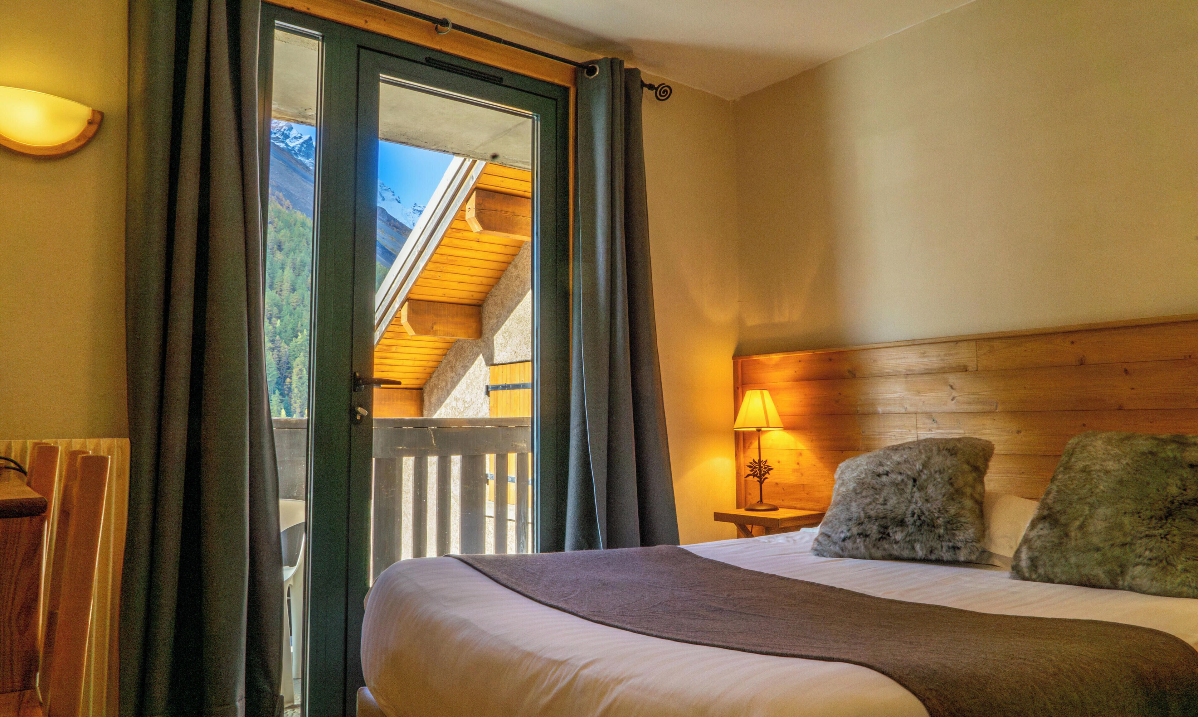 A bedroom with a bed, a nightstand with a lamp, a window with curtains, and a view of a wooden structure outside.