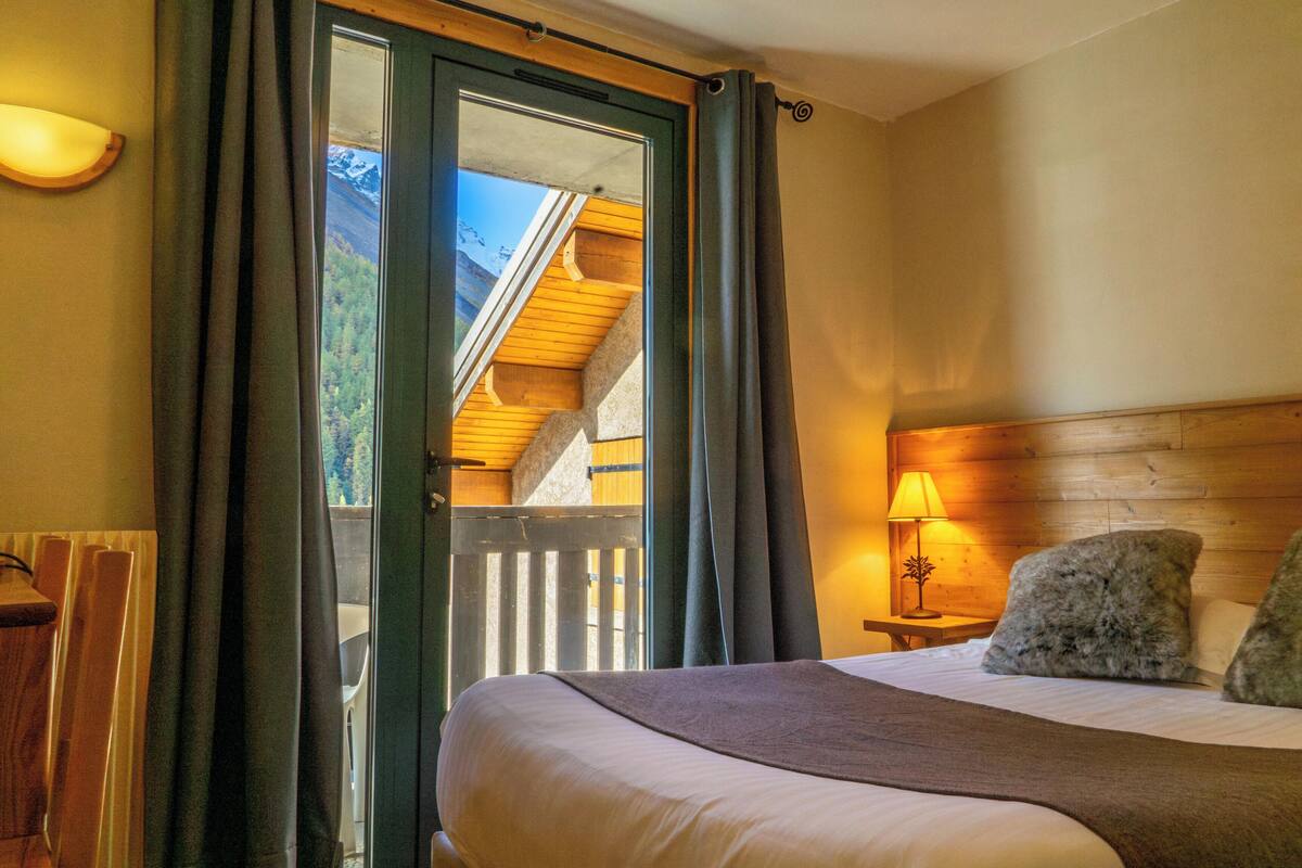 A bedroom with a bed, a nightstand with a lamp, a window with curtains, and a view of a wooden structure outside.
