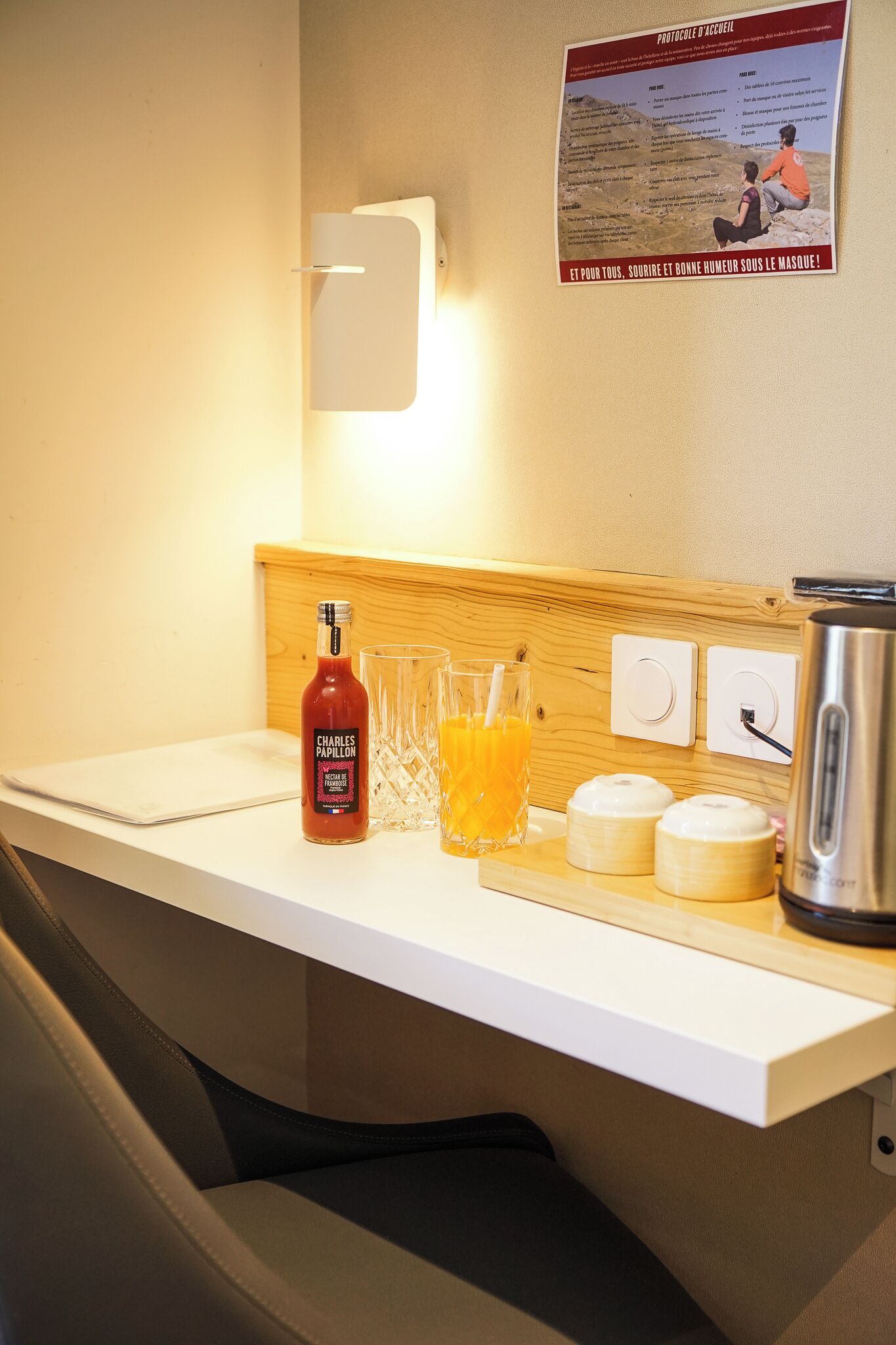 A hotel reception area with a counter, a bottle of Charles Papillon, two glasses of orange juice, and a poster on the wall.