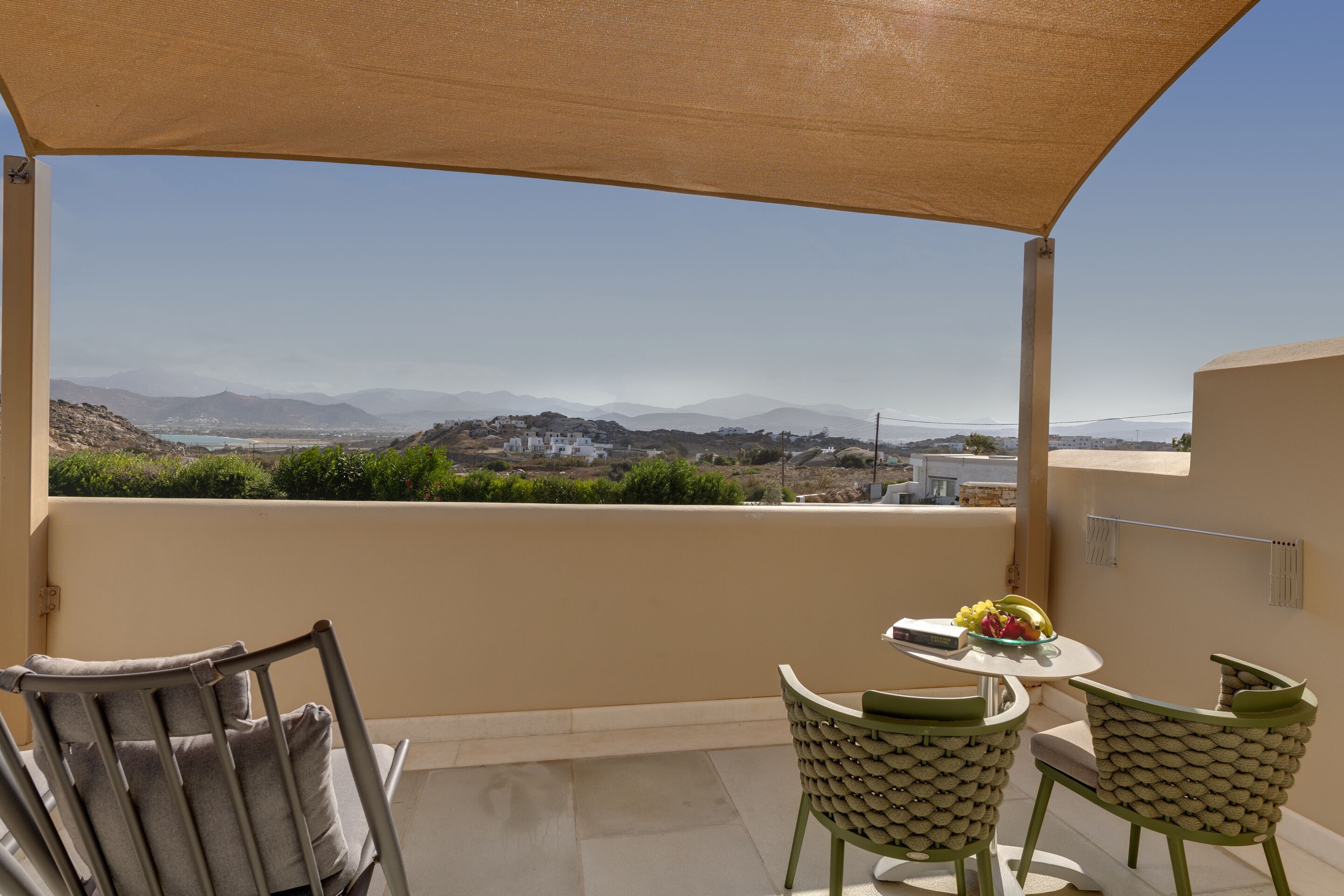 A balcony with a view of a mountainous landscape, a table with a fruit bowl, and two chairs.