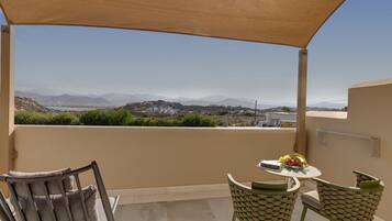 A balcony with a view of a mountainous landscape, a table with a fruit bowl, and two chairs.